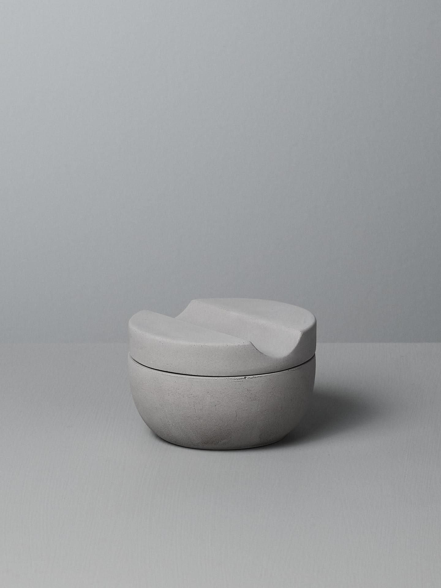 An Iris Hantverk Shaving Cup with Cedarwood Shaving Soap, a minimalist, grey ceramic cup with a curved lid, rests on a soft concrete surface, perfect for holding your cedarwood shaving soap.