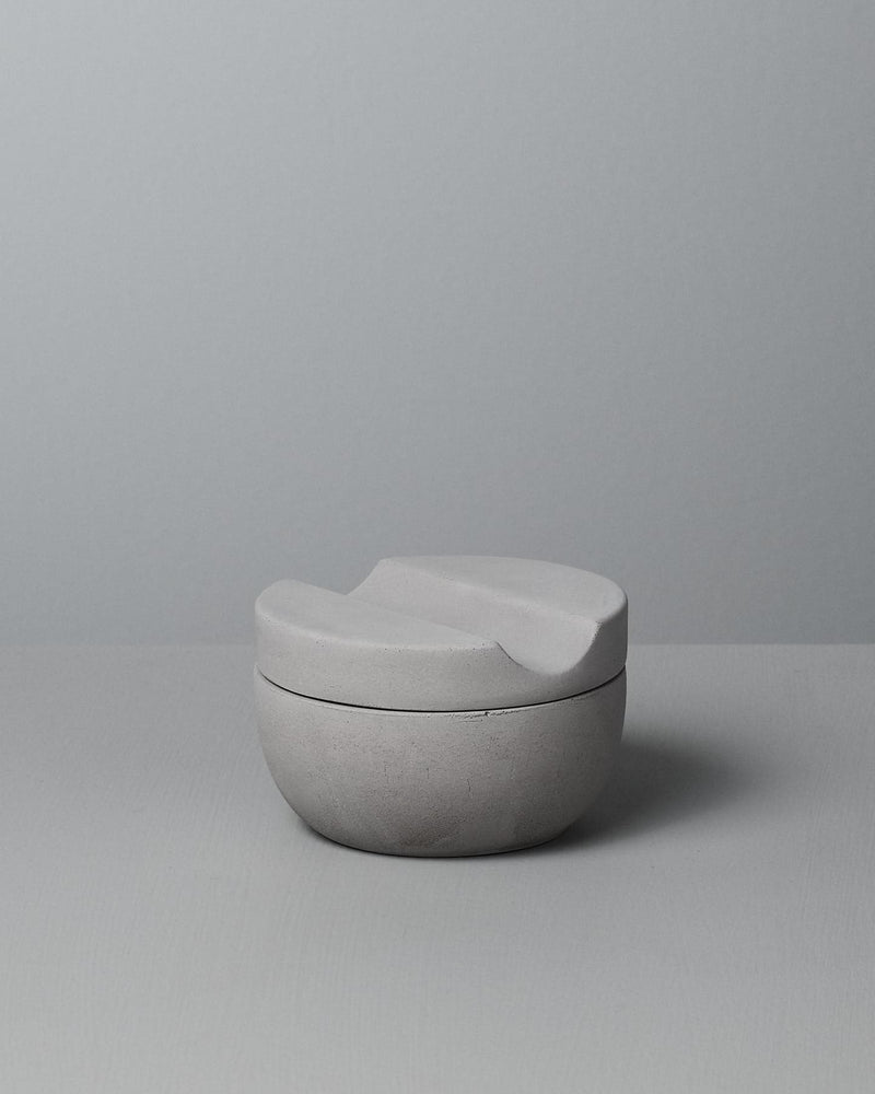 An Iris Hantverk Shaving Cup with Cedarwood Shaving Soap, a minimalist, grey ceramic cup with a curved lid, rests on a soft concrete surface, perfect for holding your cedarwood shaving soap.