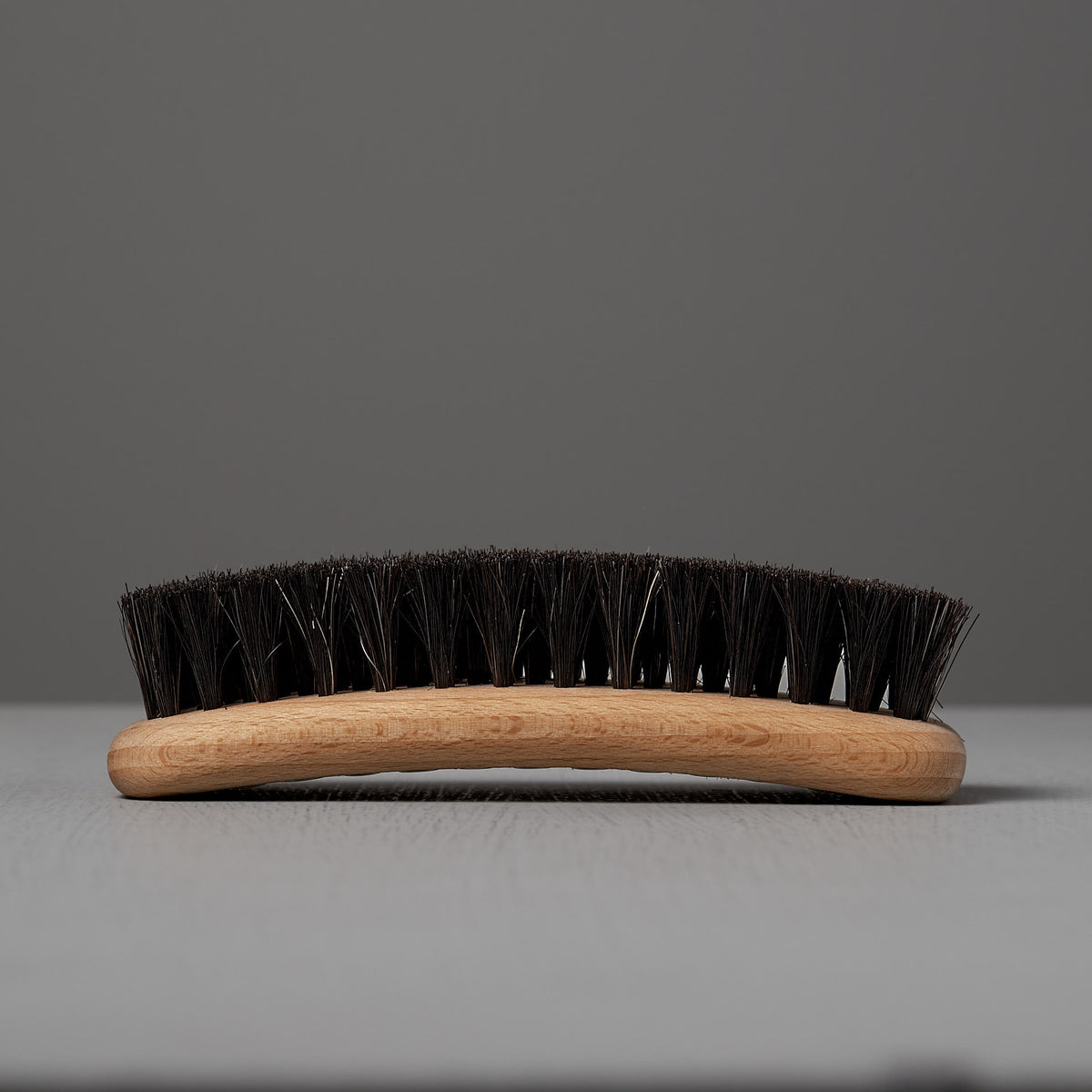 A close-up of the Iris Hantverk Shoe Brush, crafted from beech wood with dark horse hair bristles, is laid horizontally on a light-grey surface against a grey backdrop.