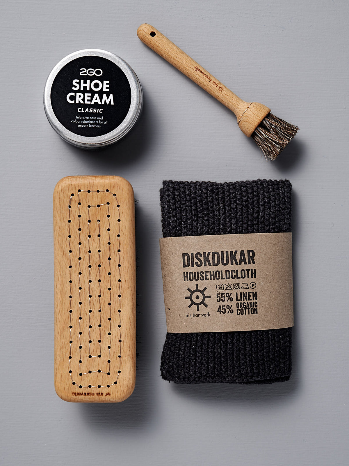 An Iris Hantverk Shoe Care Kit, featuring a wooden shoe brush, a black shoe polish tin, and a folded household cloth, is neatly arranged on a gray surface.