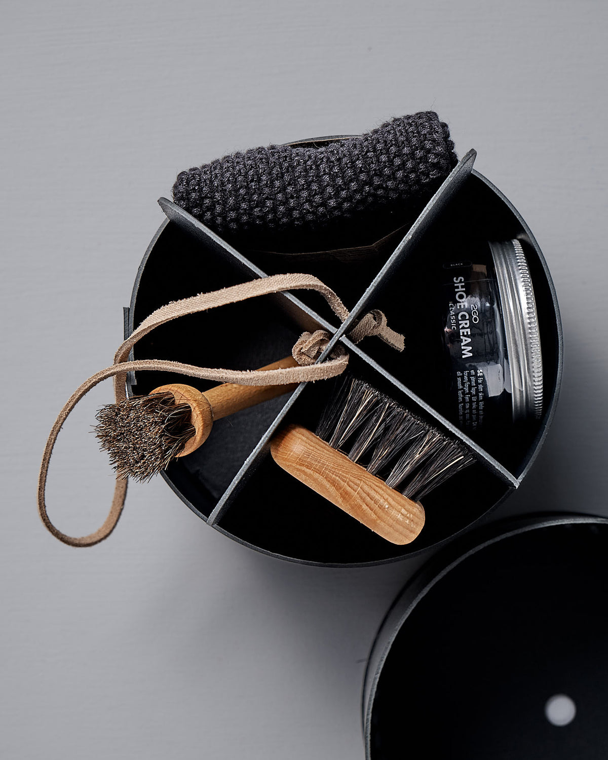 The Iris Hantverk Shoe Care Kit comes in a circular box and includes essential tools for maintaining your footwear's shine and longevity: a shoe brush, polishing cloth, jar of black shoe polish, and polishing pad.