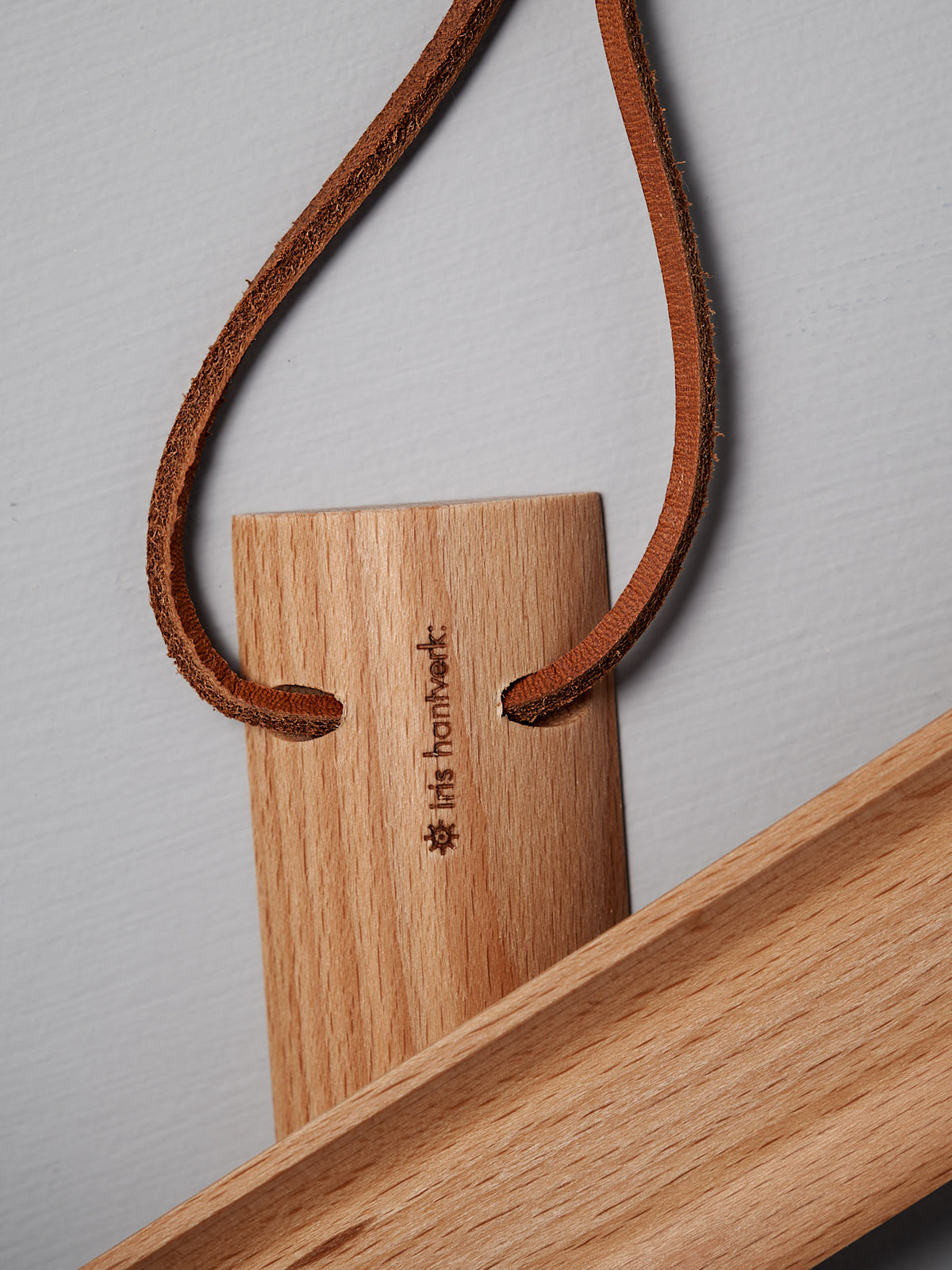 A short shoe horn crafted from oiled beech wood, featuring carved characters and attached to a leather strap, sits gracefully on a light gray surface, invoking the essence of Iris Hantverk&#39;s design.