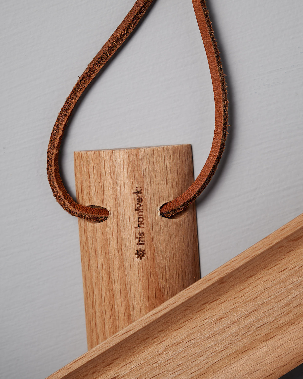 A short shoe horn crafted from oiled beech wood, featuring carved characters and attached to a leather strap, sits gracefully on a light gray surface, invoking the essence of Iris Hantverk's design.