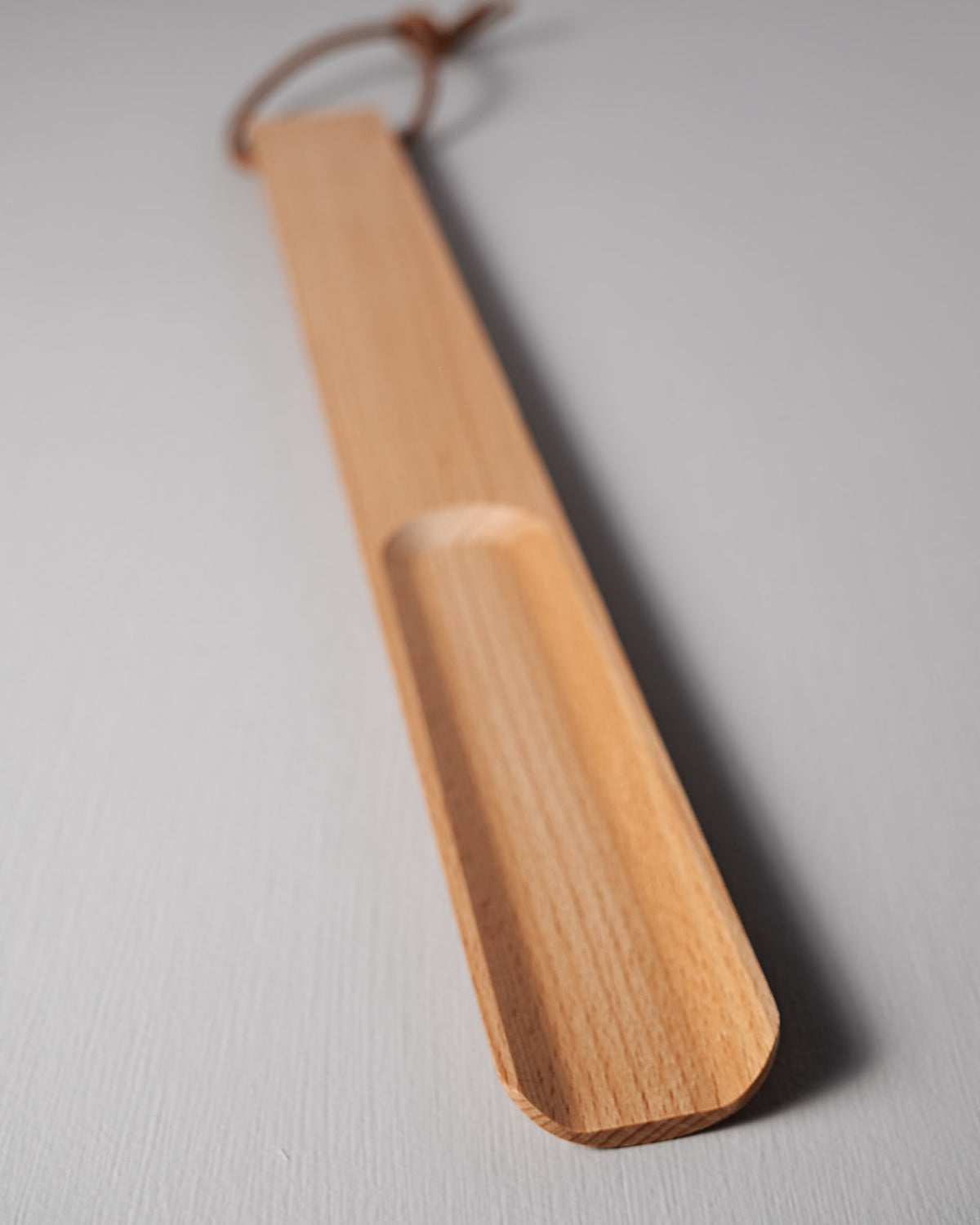 A long shoehorn from Iris Hantverk, crafted from oiled beech wood, boasts a smooth finish and includes a leather loop at one end, elegantly displayed on a light gray surface.