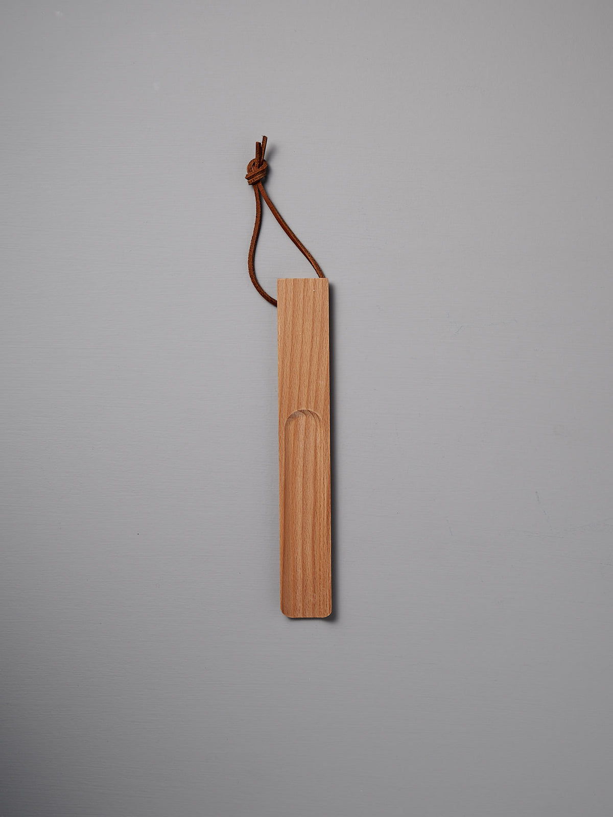 A beautifully crafted shoe horn made of oiled beech, featuring a leather loop at the top, rests elegantly on a gray surface. Brought to you by the renowned brand Iris Hantverk.