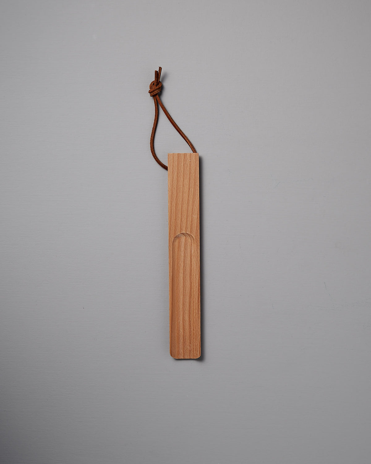 A beautifully crafted shoe horn made of oiled beech, featuring a leather loop at the top, rests elegantly on a gray surface. Brought to you by the renowned brand Iris Hantverk.