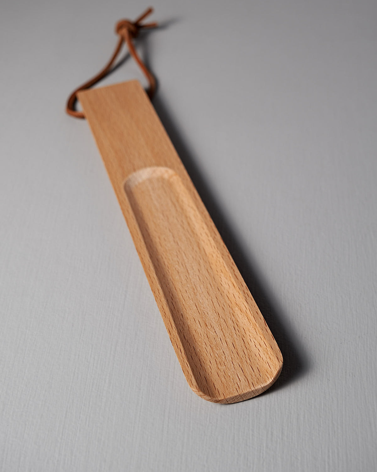 An Iris Hantverk Shoe Horn – Short ⋄ Long, made from oiled beech and featuring a leather loop, rests gracefully on a light gray surface.