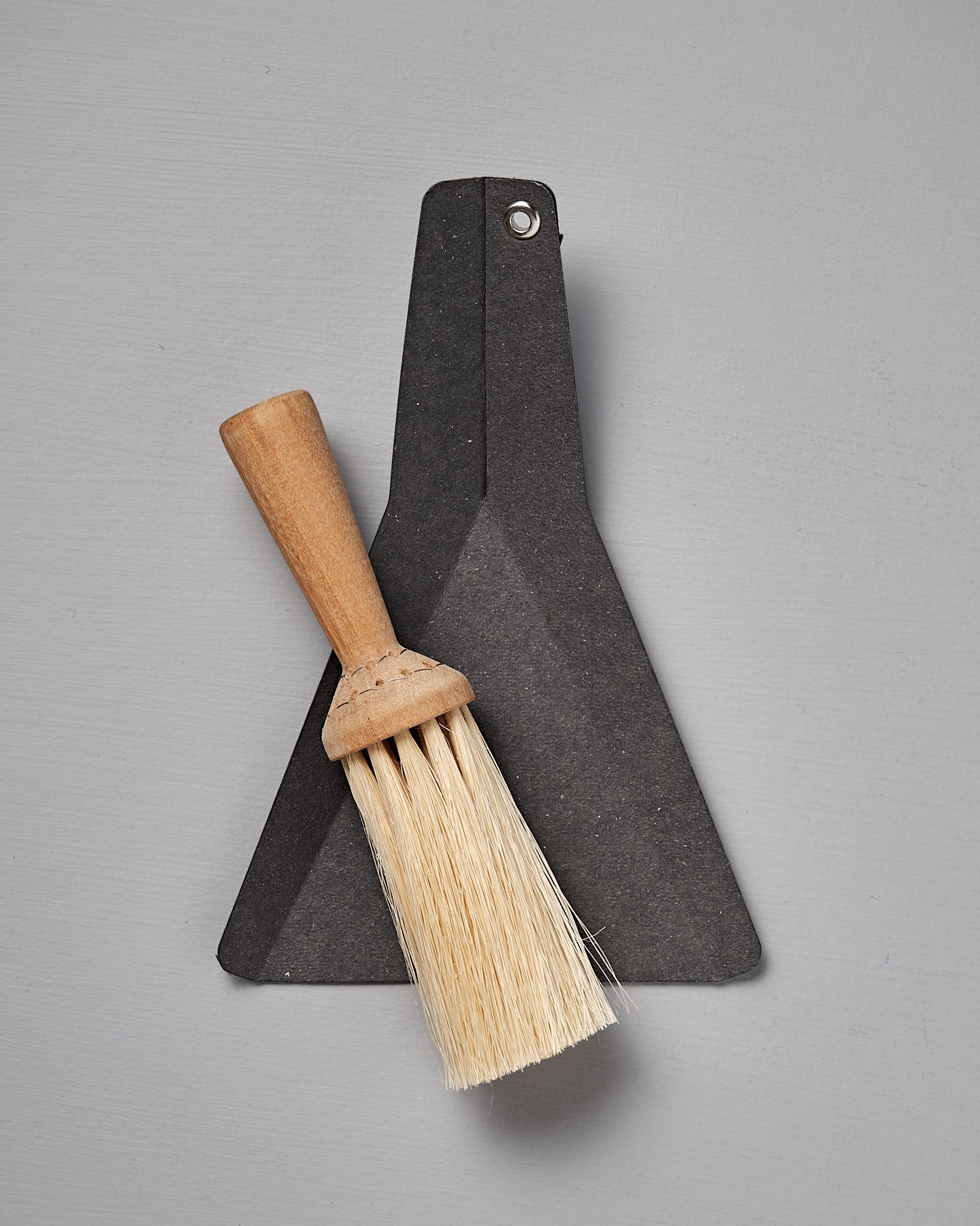 The Iris Hantverk Table Broom & Shovel Set features a small hand brush with an oiled birch handle and tampico bristles, paired with a flat black dustpan, shown against a gray background.