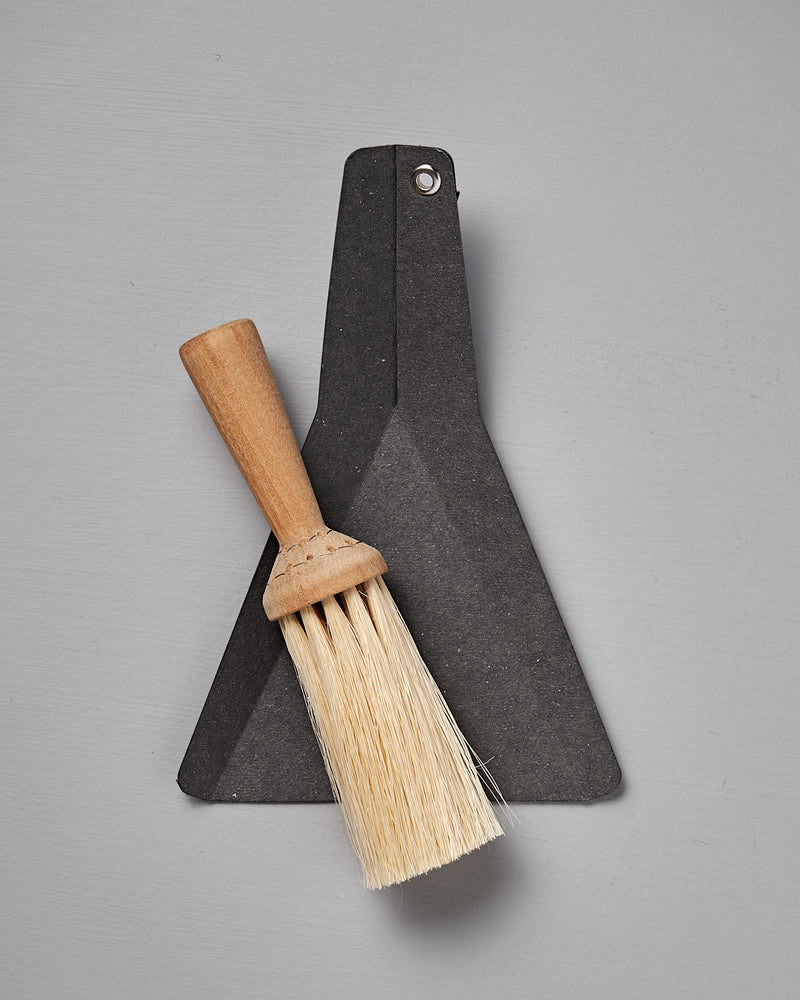 The Iris Hantverk Table Broom & Shovel Set features a small hand brush with an oiled birch handle and tampico bristles, paired with a flat black dustpan, shown against a gray background.