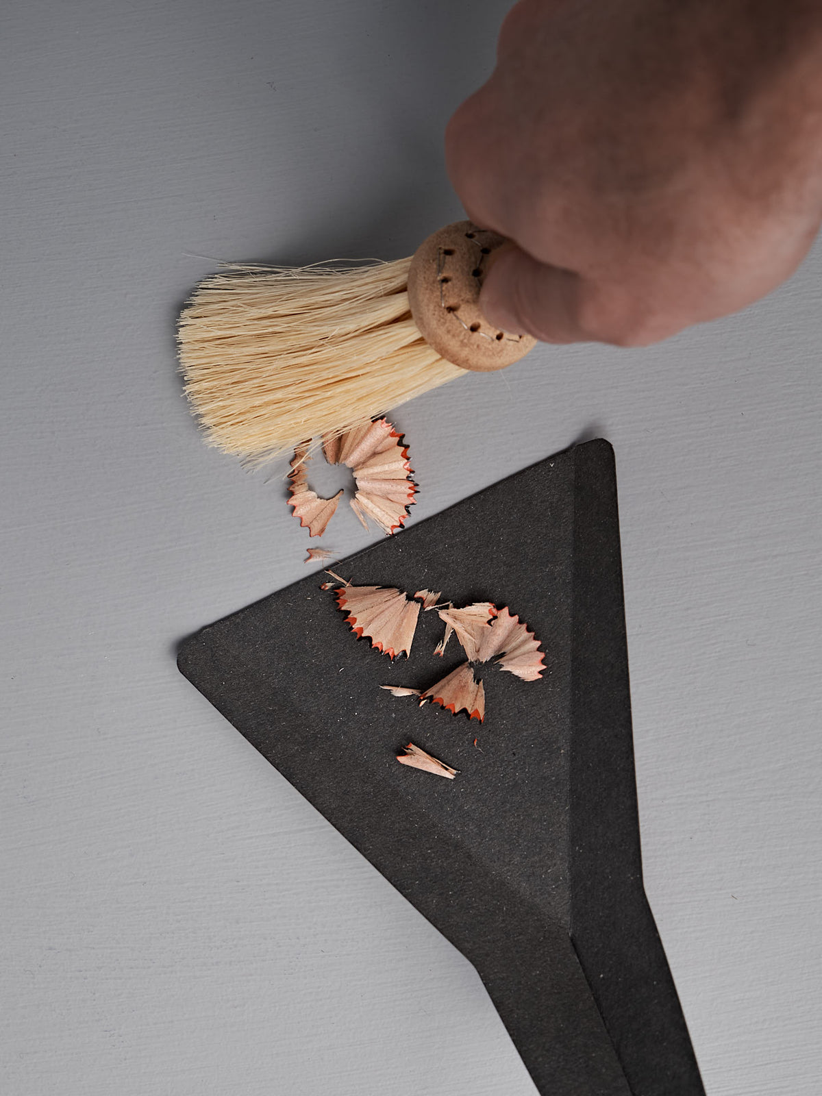A hand sweeps pencil shavings onto a black dustpan using the Iris Hantverk Table Broom &amp; Shovel Set with tampico fiber bristles on a gray surface.