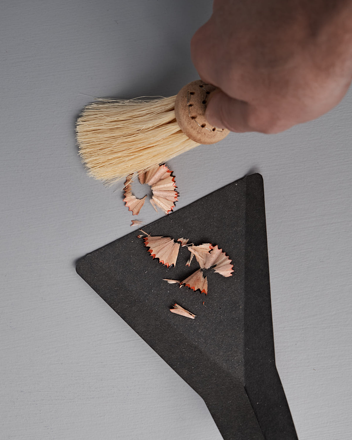 A hand sweeps pencil shavings onto a black dustpan using the Iris Hantverk Table Broom & Shovel Set with tampico fiber bristles on a gray surface.