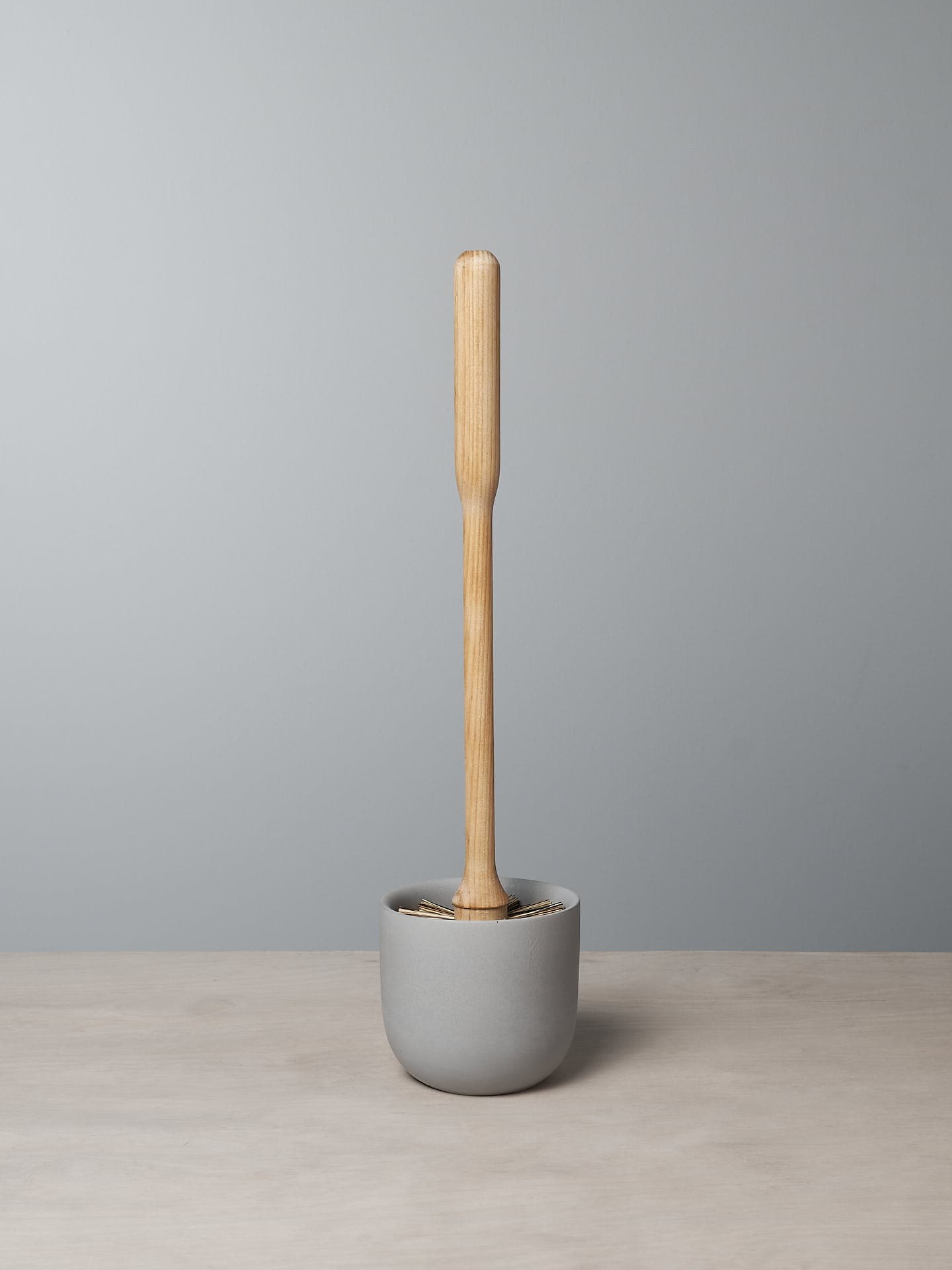 An Iris Hantverk Toilet Brush with Concrete Cup stands upright in a gray polymer mix concrete holder on a light wood surface against a plain gray wall.