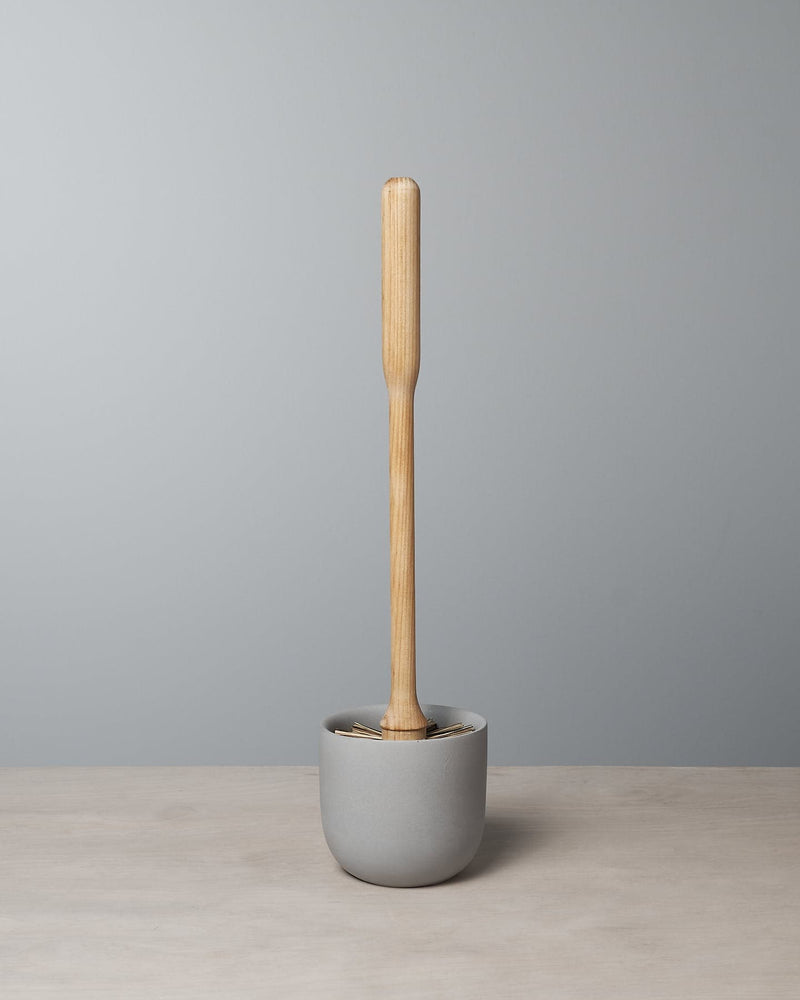 An Iris Hantverk Toilet Brush with Concrete Cup stands upright in a gray polymer mix concrete holder on a light wood surface against a plain gray wall.