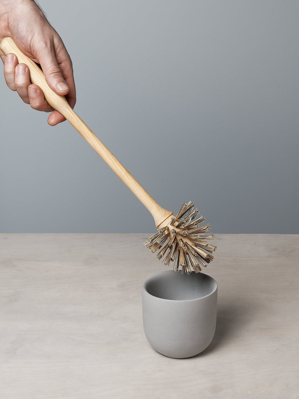 A hand holding an Iris Hantverk Toilet Brush + Toilet Roll Holder Set with a wooden handle and bristles is positioned above a small, grey cup on a light-colored surface.