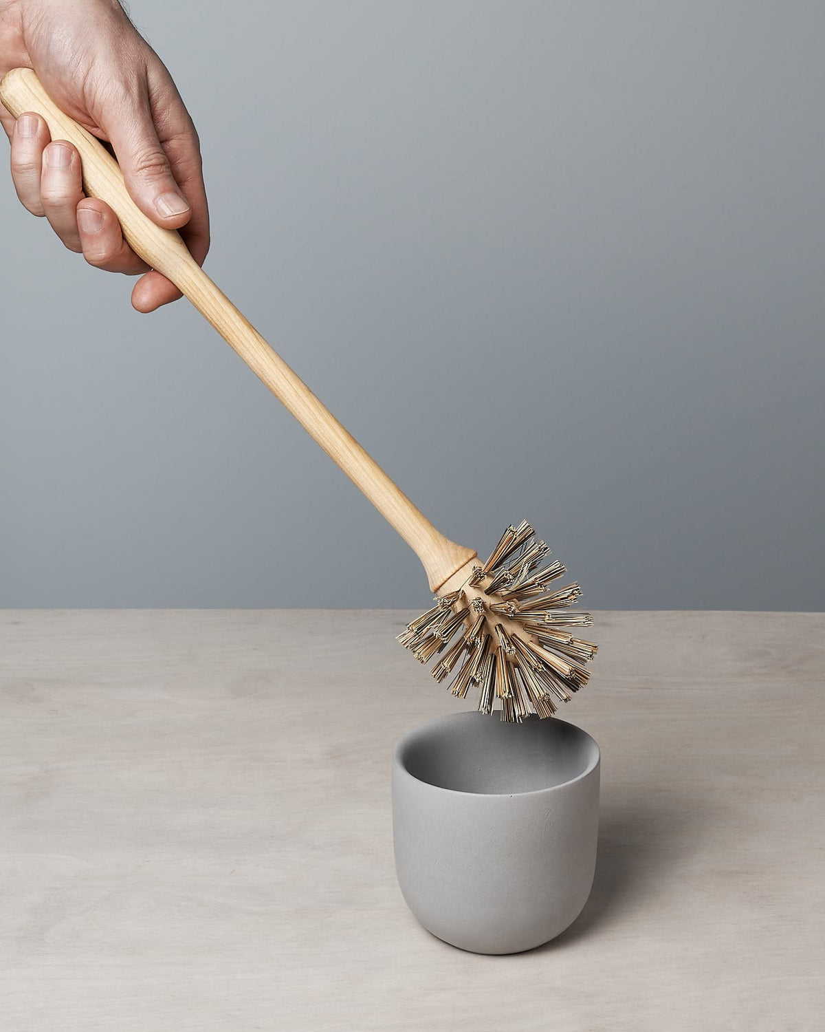 A hand holds an Iris Hantverk Toilet Brush & Toilet Roll Holder over a gray cup on a light wooden surface, highlighting the elegance of its minimalistic design.