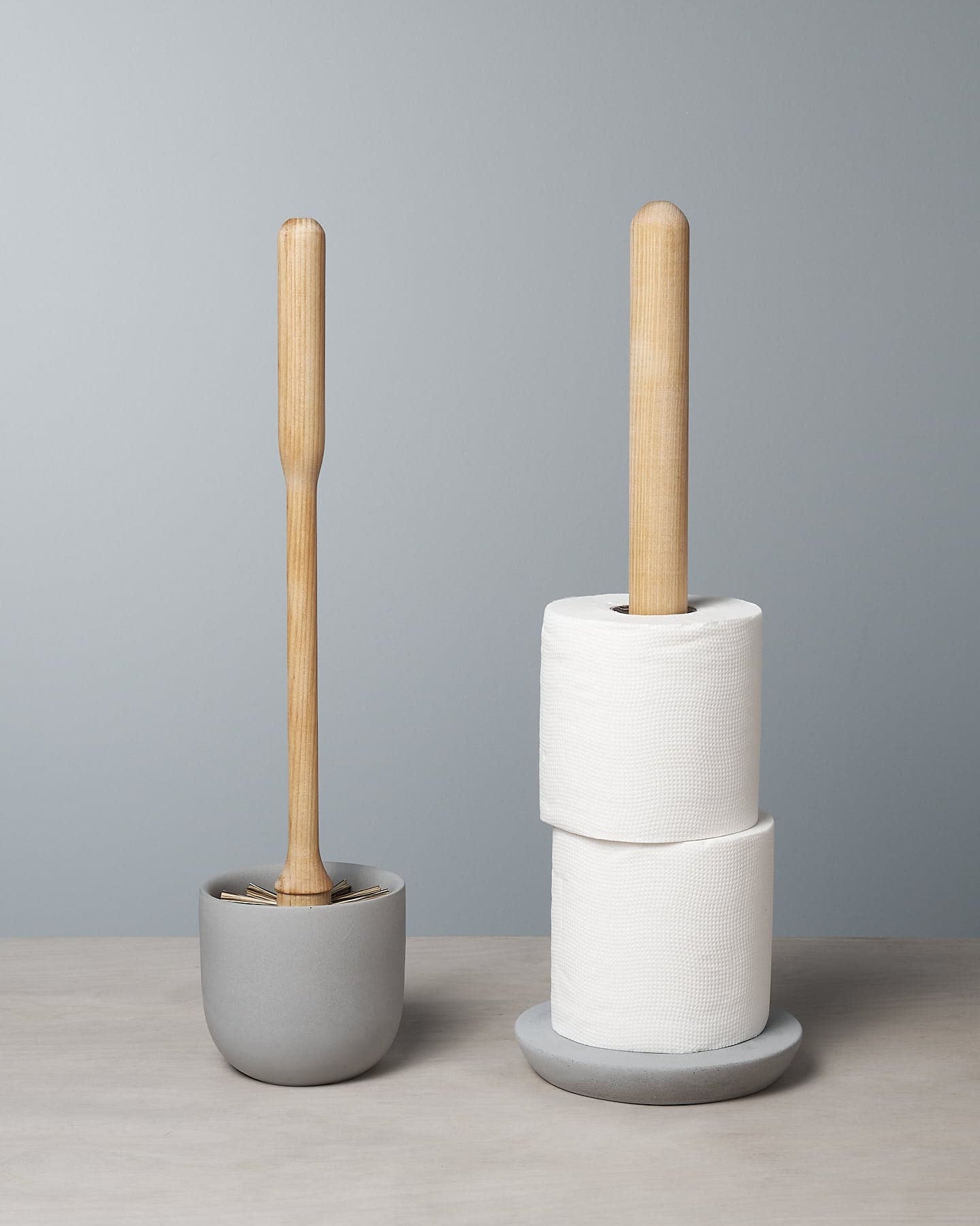 The Toilet Brush & Toilet Roll Holder – Bundle by Iris Hantverk, featuring an elegantly crafted toilet brush and accompanying wooden-handled toilet roll holder with two rolls, is beautifully displayed on a soft concrete surface in soothing light gray tones.