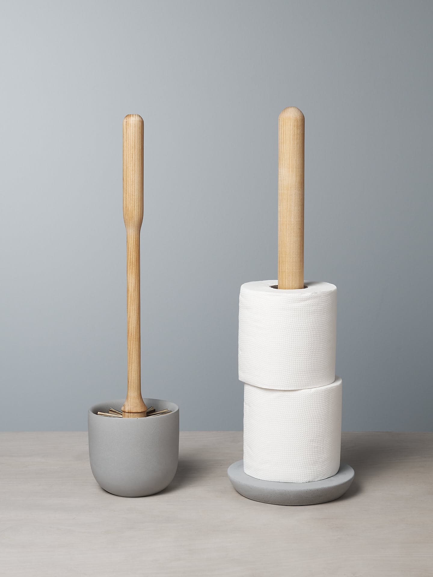 The Toilet Brush & Toilet Roll Holder – Bundle by Iris Hantverk, featuring an elegantly crafted toilet brush and accompanying wooden-handled toilet roll holder with two rolls, is beautifully displayed on a soft concrete surface in soothing light gray tones.