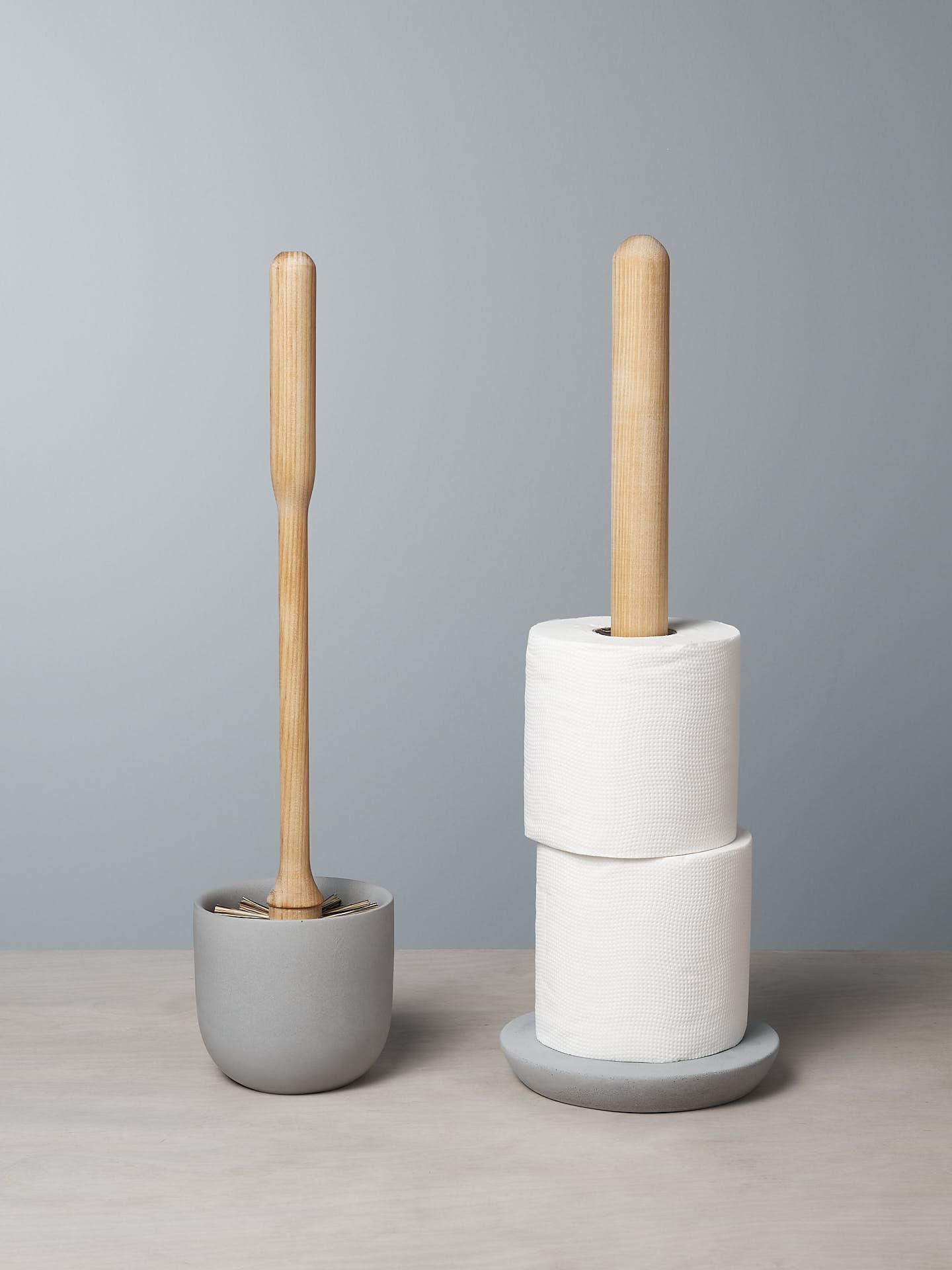 An Iris Hantverk Toilet Brush + Toilet Roll Holder Set is placed next to a toilet roll holder containing two rolls of white toilet paper on a soft concrete surface.