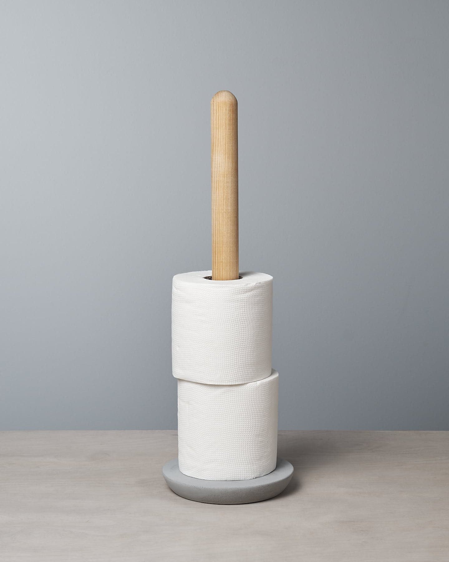 An Iris Hantverk Toilet Roll Holder with two rolls of toilet paper stacked on it, standing on a soft concrete surface against a light gray background.