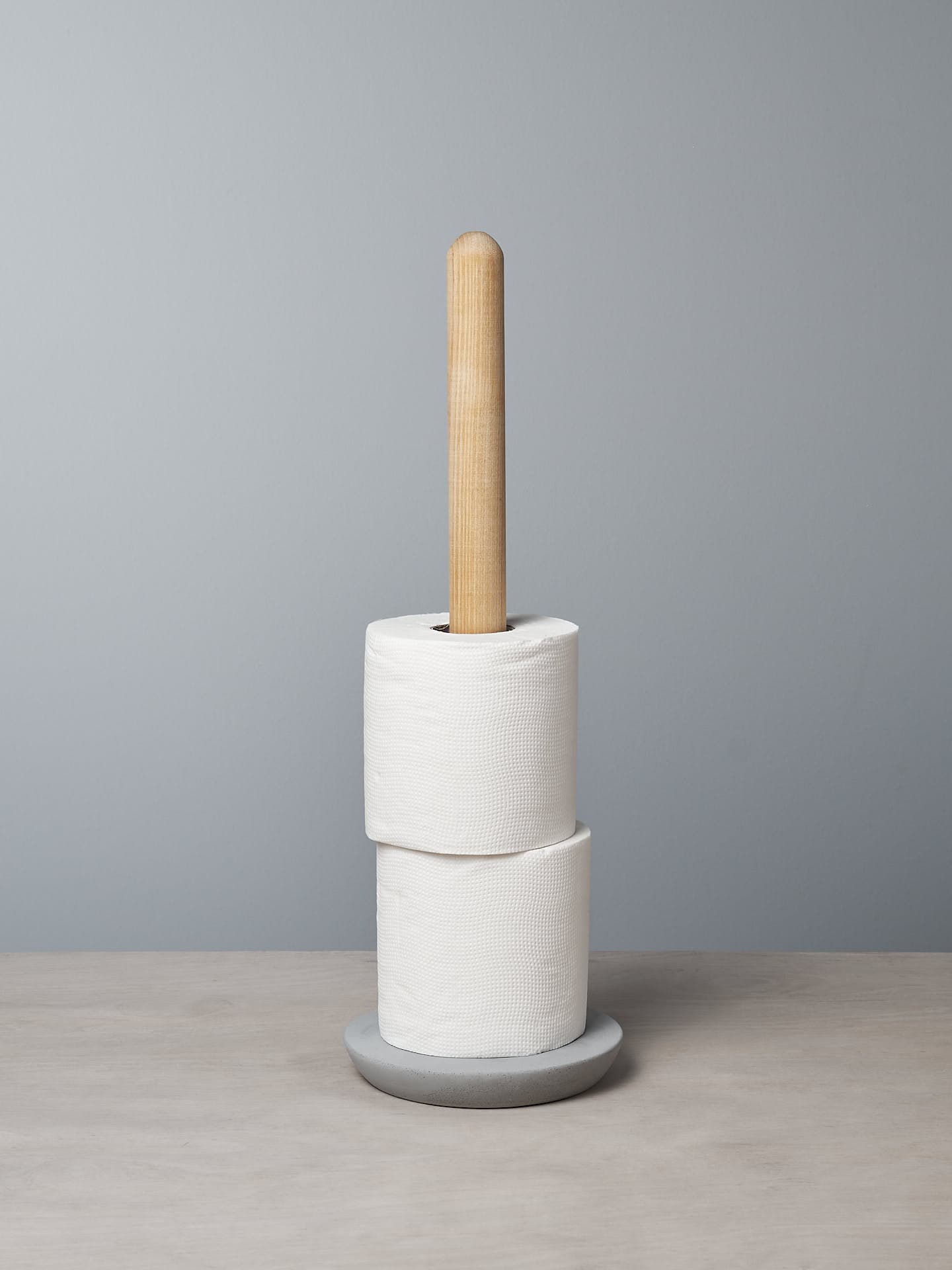 An Iris Hantverk Toilet Roll Holder with two rolls of toilet paper stacked on it, standing on a soft concrete surface against a light gray background.
