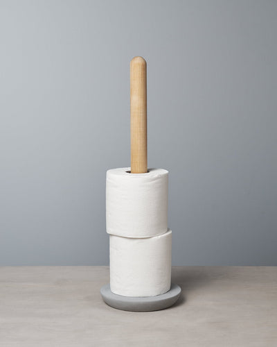 An Iris Hantverk Toilet Roll Holder with two rolls of toilet paper stacked on it, standing on a soft concrete surface against a light gray background.