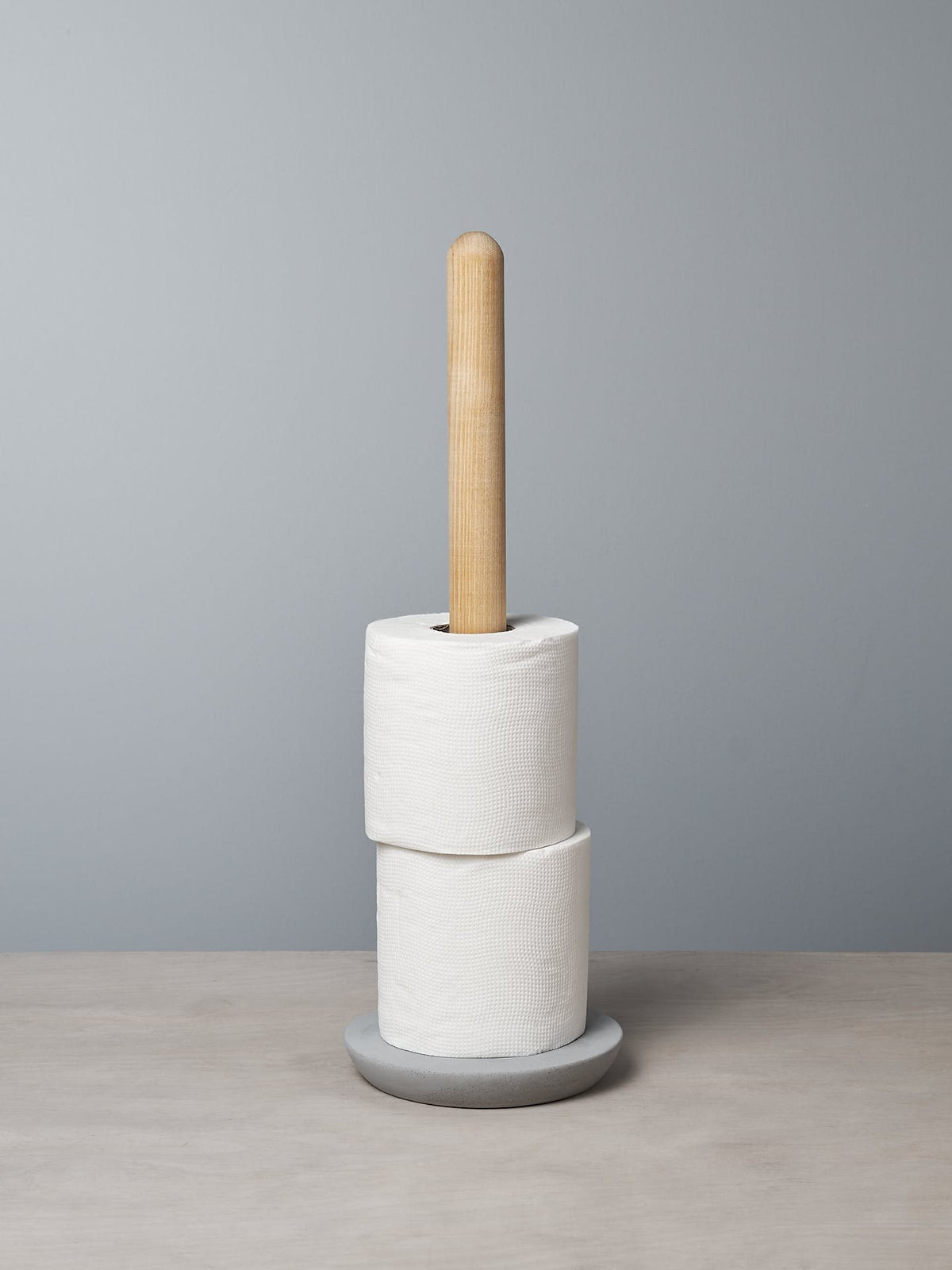 An Iris Hantverk Toilet Brush + Toilet Roll Holder Set stands elegantly on a gray background, holding two rolls of toilet paper.