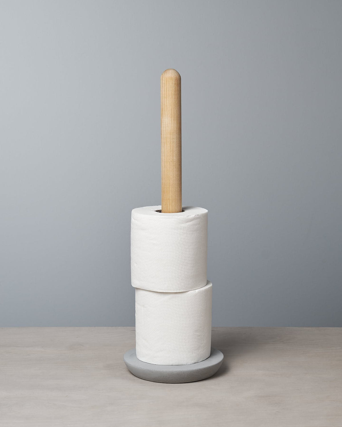 A pair of white toilet paper rolls are neatly positioned on the stylish toilet roll holder with a gray base from the Iris Hantverk Toilet Brush & Toilet Roll Holder – Bundle, set against a simple gray wall.