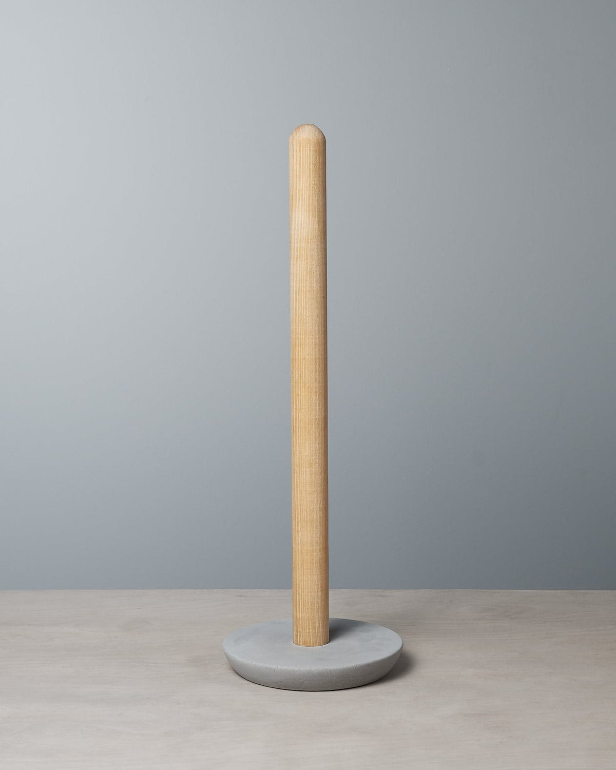 An Iris Hantverk Toilet Roll Holder crafted from beechwood with a soft concrete base stands on a light gray surface against a pale blue background.