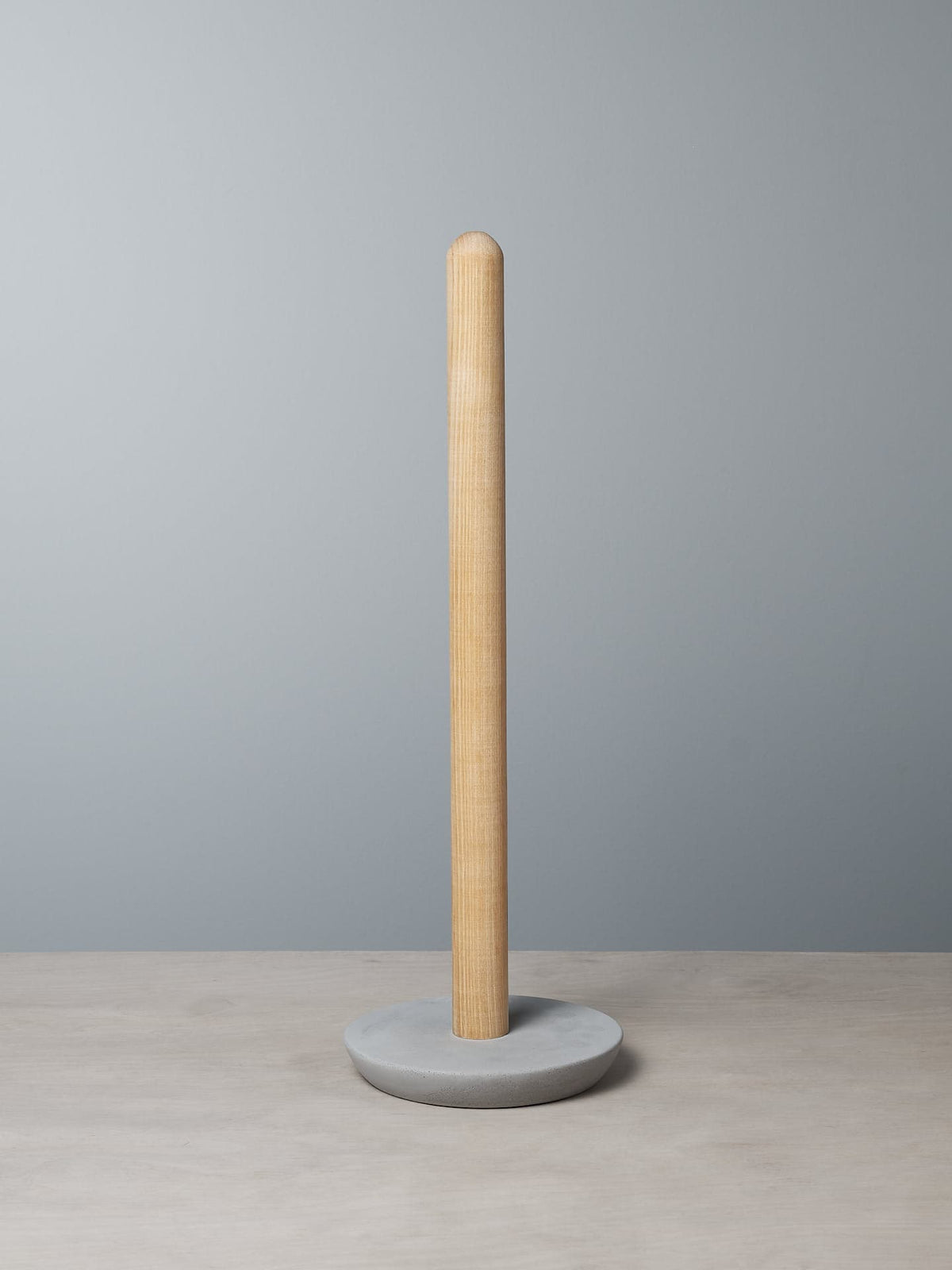 An Iris Hantverk Toilet Brush + Toilet Roll Holder Set with a cylindrical base stands on a soft concrete surface against a gray background.