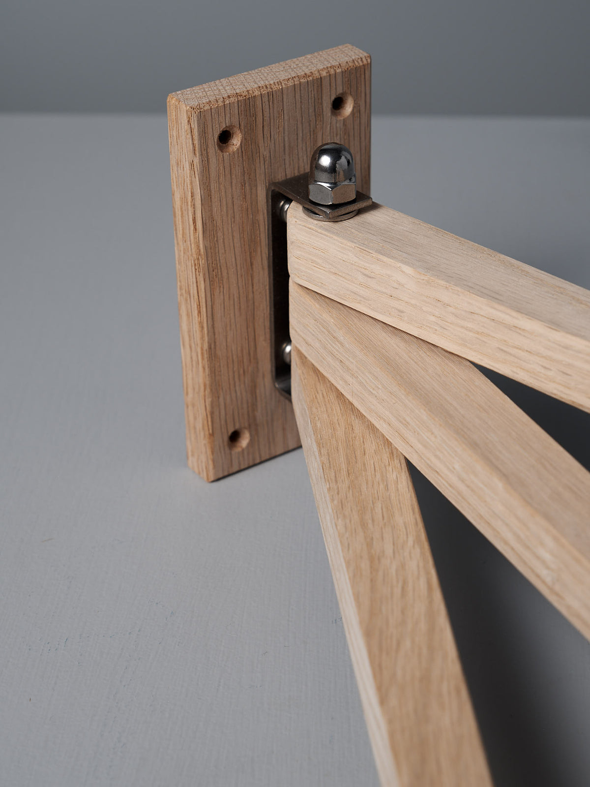 A close-up of a wooden joint with a metal hinge connecting two oak pieces highlights the elegance of the Oak Towel Drying Rack – 3 Arm by Iris Hantverk, set against a gray bathroom surface.