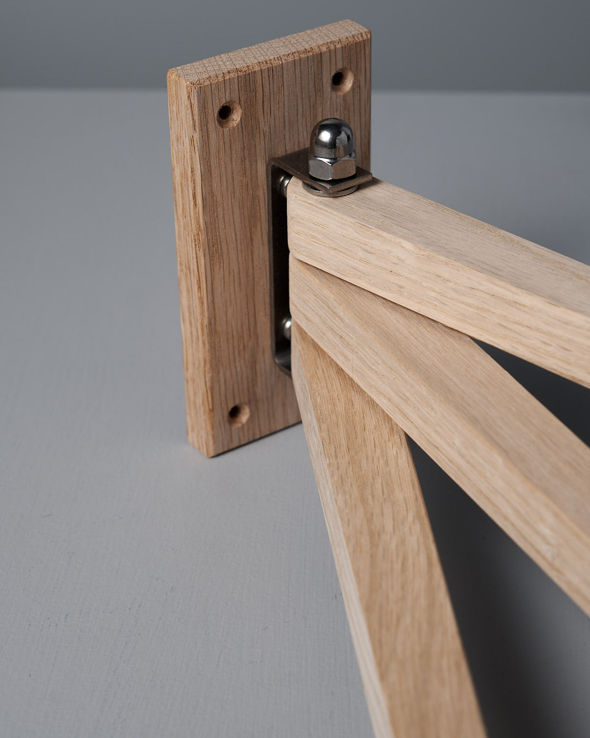 A close-up of a wooden joint with a metal hinge connecting two oak pieces highlights the elegance of the Oak Towel Drying Rack – 3 Arm by Iris Hantverk, set against a gray bathroom surface.
