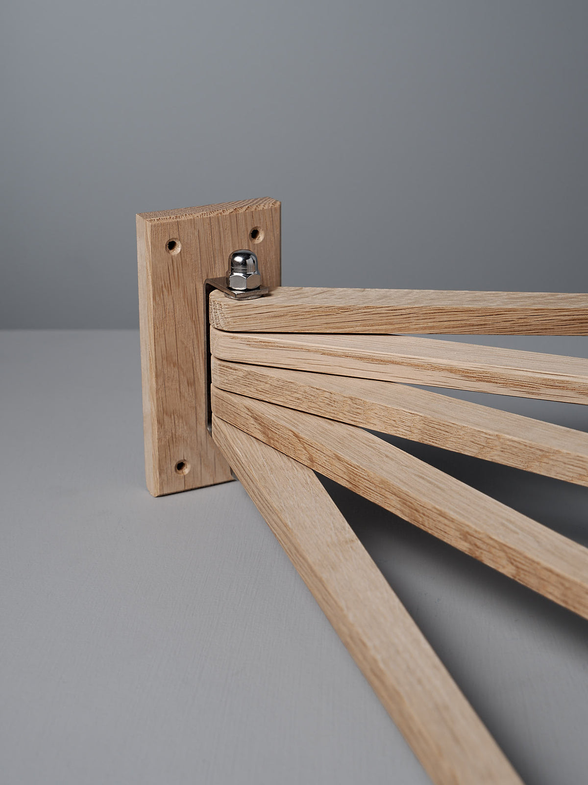 The Oak Towel Drying Rack – 5 Arm by Iris Hantverk is a wooden mechanical structure crafted from solid oak, featuring foldable arms that pivot from a central joint on a square base, set against a gray background.