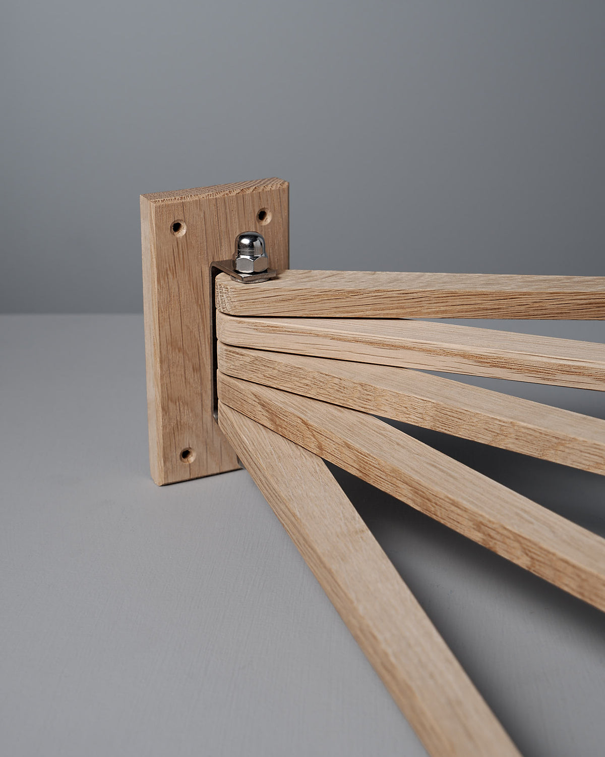 The Oak Towel Drying Rack – 5 Arm by Iris Hantverk is a wooden mechanical structure crafted from solid oak, featuring foldable arms that pivot from a central joint on a square base, set against a gray background.