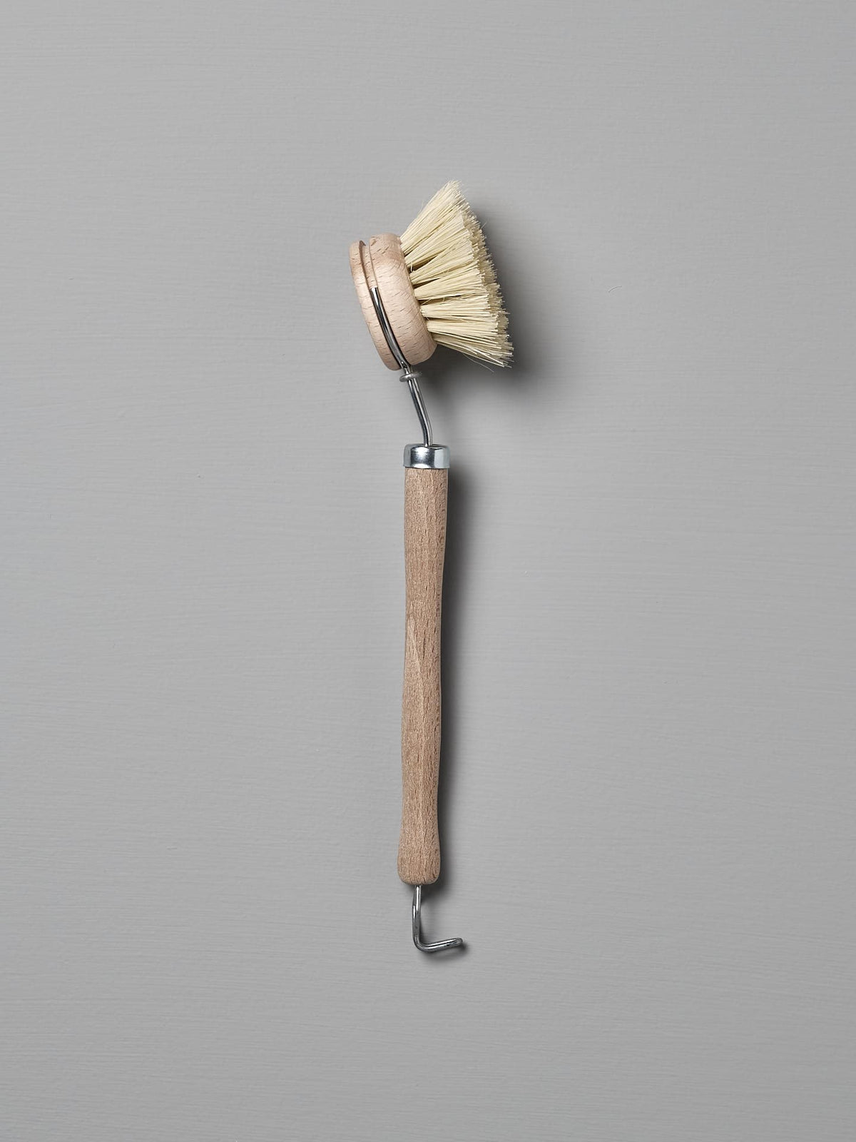 Iris Hantverk&#39;s Dish Brush with Replaceable Head, featuring a rounded head and stiff Tampico fibre bristles, along with a long handle and metal hook at the end, placed against a grey background.