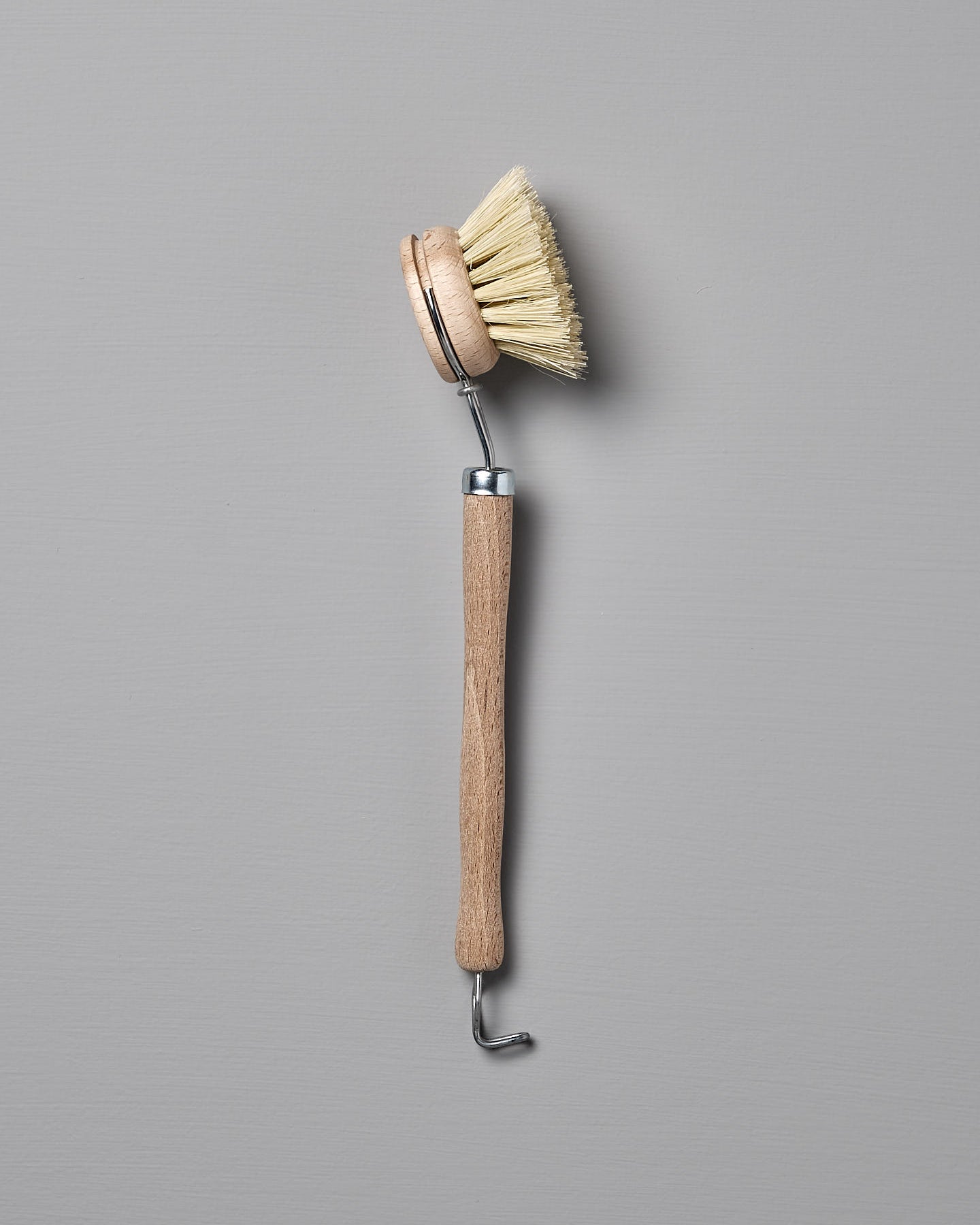 Iris Hantverk's Dish Brush with Replaceable Head, featuring a rounded head and stiff Tampico fibre bristles, along with a long handle and metal hook at the end, placed against a grey background.