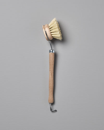 Iris Hantverk's Dish Brush with Replaceable Head, featuring a rounded head and stiff Tampico fibre bristles, along with a long handle and metal hook at the end, placed against a grey background.