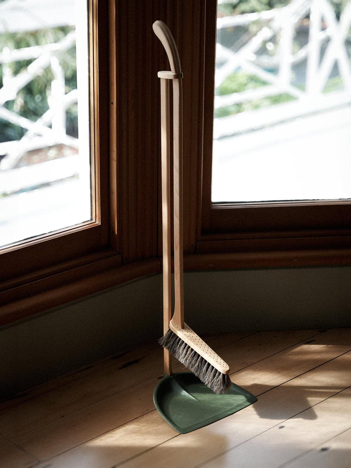 An Iris Hantverk Long Handled Dustpan &amp; Brush Set – Moss Green stands upright positioned against a wooden windowsill, with sunlight streaming through.