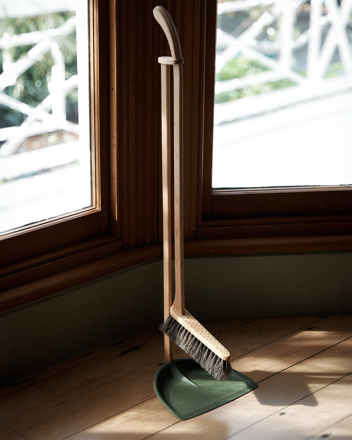 An Iris Hantverk Long Handled Dustpan & Brush Set – Moss Green stands upright positioned against a wooden windowsill, with sunlight streaming through.