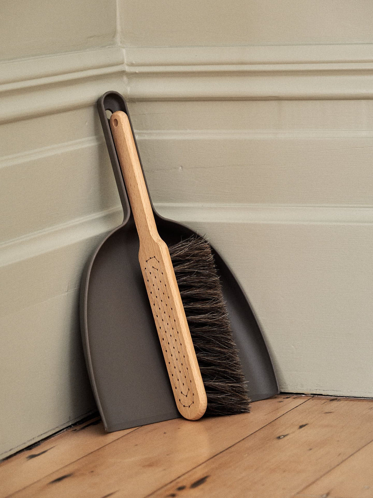 A Dustpan &amp; Brush Set – Umber from Iris Hantverk with a beech wood handle and horsehair brush rests inside a bio-polyethylene dustpan, both leaning against a corner of a room with wooden flooring and painted walls.