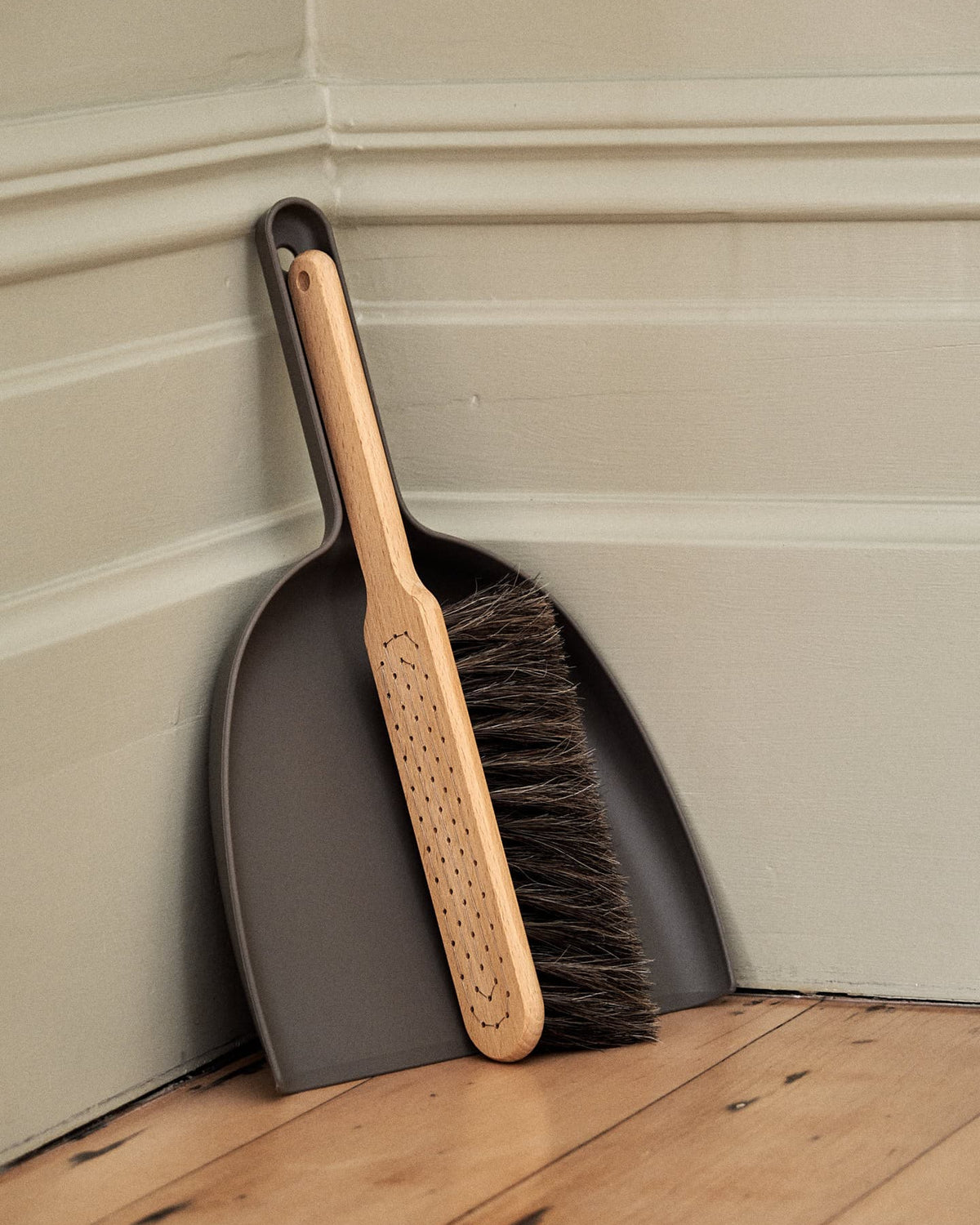 A Dustpan & Brush Set – Umber from Iris Hantverk with a beech wood handle and horsehair brush rests inside a bio-polyethylene dustpan, both leaning against a corner of a room with wooden flooring and painted walls.