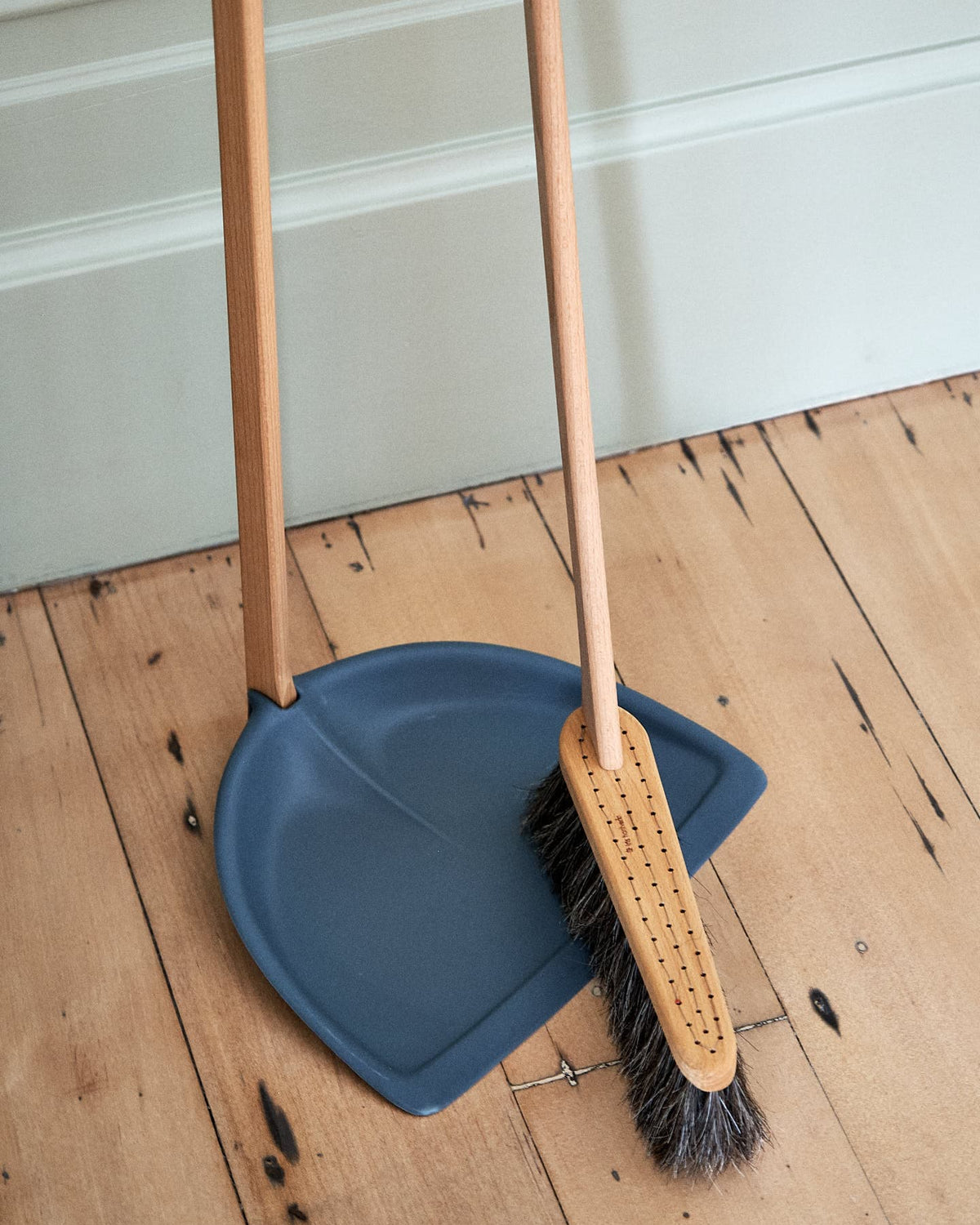 A Long Handled Dustpan & Brush Set – Petrol Blue by Iris Hantverk with wooden handles are placed on a wooden floor next to a light green wall, crafted from eco-friendly materials.
