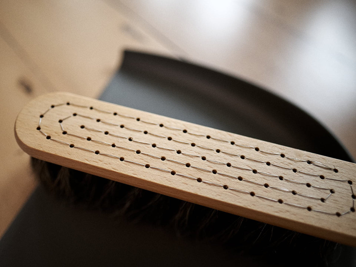 A close-up of an Iris Hantverk Dustpan &amp; Brush Set – Umber with bristles, placed on the edge of a black dustpan. The brush, crafted from oiled beech wood, features rows of evenly spaced holes on its handle.