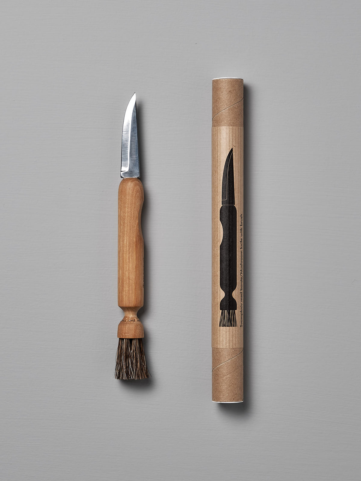 The Iris Hantverk Mushroom Brush &amp; Knife, featuring a wooden handle and horsehair brush, is shown beside its cylindrical cardboard package with an illustration of the product.