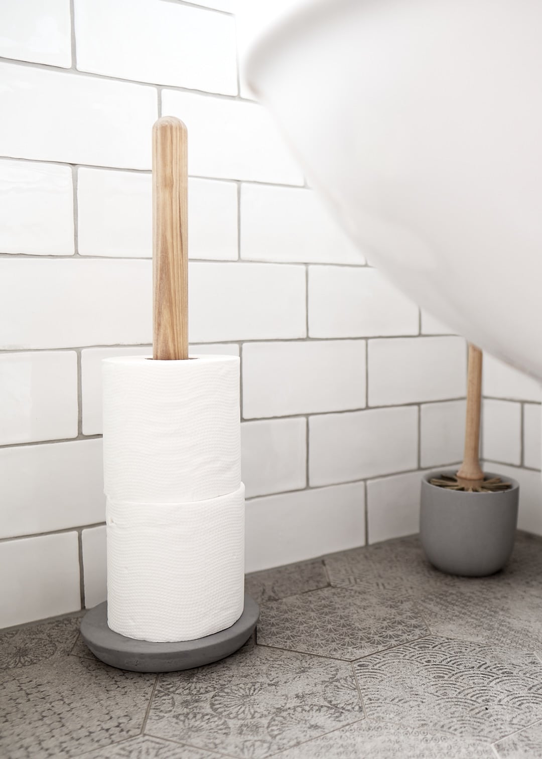 A roll of toilet paper on an Iris Hantverk Toilet Brush + Toilet Roll Holder Set next to a toilet brush in a white-tiled bathroom.