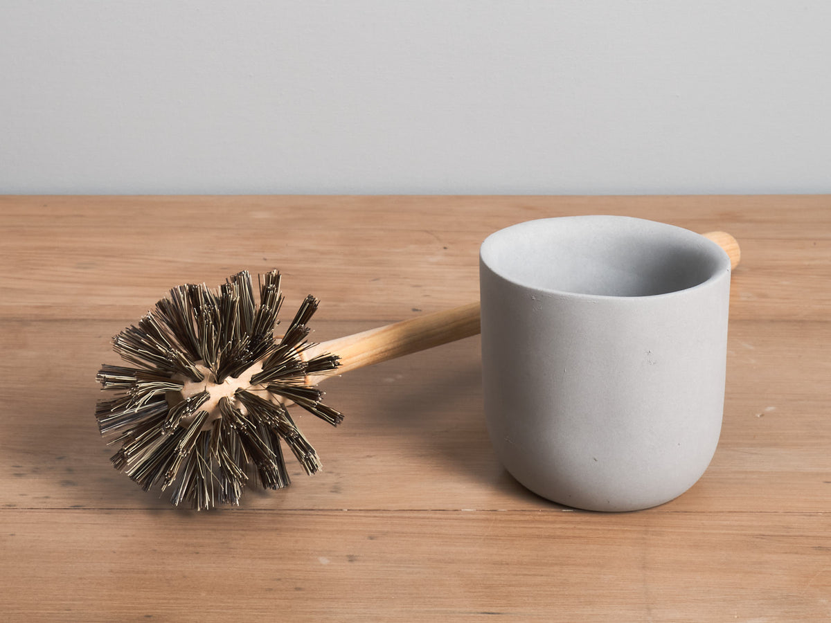 A Iris Hantverk toilet brush + toilet roll holder set and a white ceramic mug on a wooden surface.