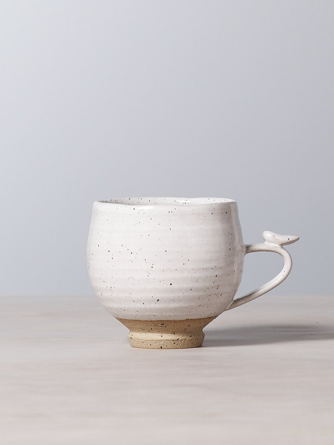 A Bird Handle Cup – White with a bird shape handle sitting on a table from Jino Ceramic Studio.