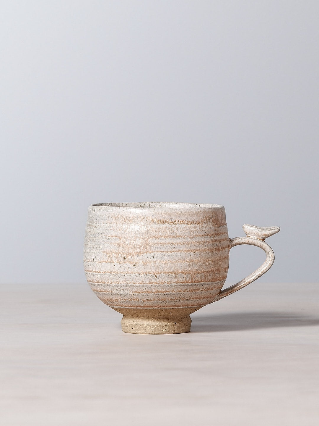 A small Bird Handle Cup – Pink Salt with a handle on a table, adorned with a graceful bloom by Jino Ceramic Studio.