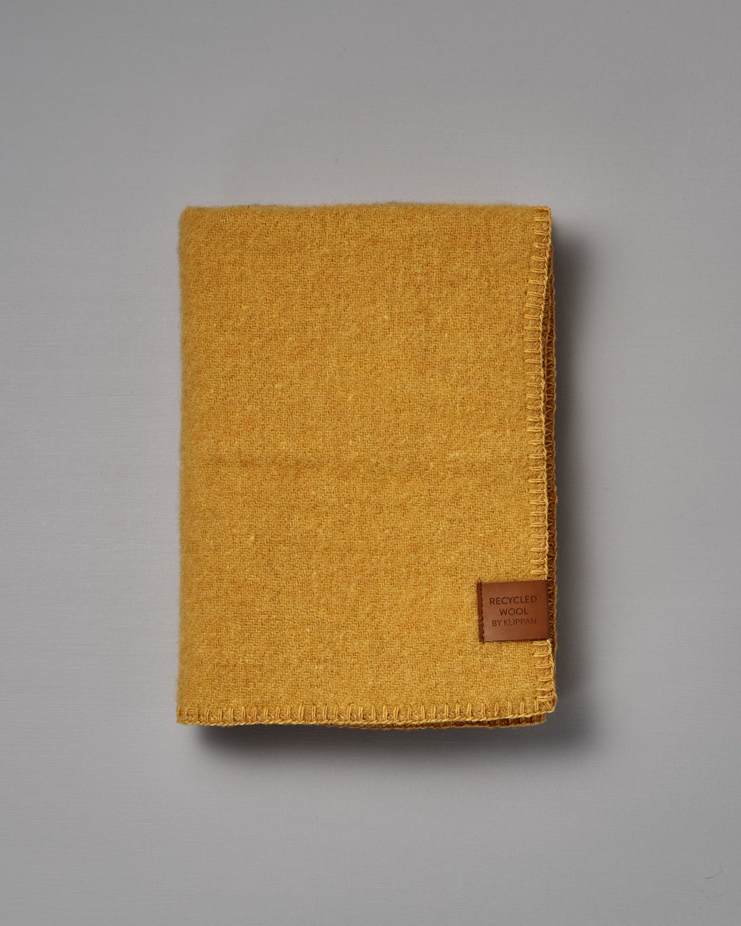 Folded True Baby Blanket – Straw, made from yellow recycled wool and featuring a brown “Recycled Wool by Re-born” label, crafted by Klippan Sweden, placed on a plain grey surface.
