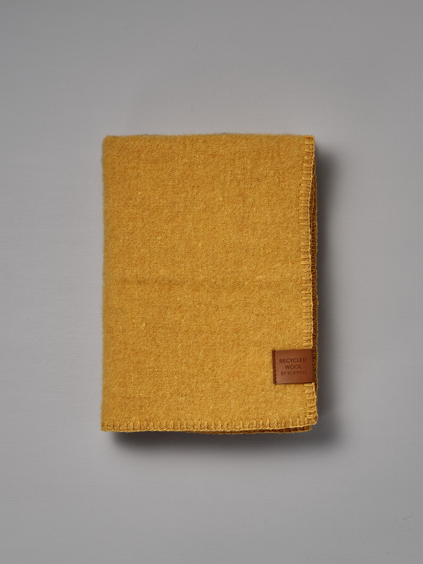 Folded True Baby Blanket – Straw, made from yellow recycled wool and featuring a brown “Recycled Wool by Re-born” label, crafted by Klippan Sweden, placed on a plain grey surface.