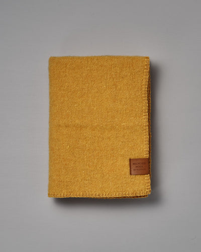Folded True Baby Blanket – Straw, made from yellow recycled wool and featuring a brown “Recycled Wool by Re-born” label, crafted by Klippan Sweden, placed on a plain grey surface.
